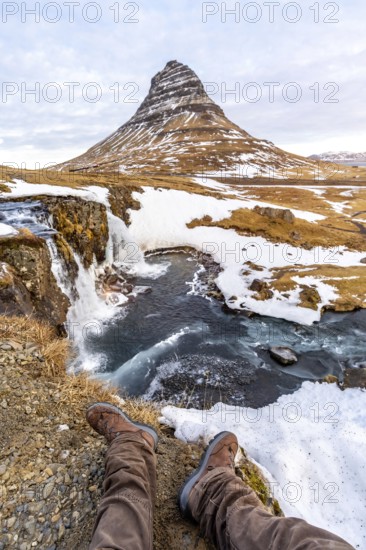 POV of a man sitting on the waterfall with snow on the famous Kirkjufell mountain in winter Iceland