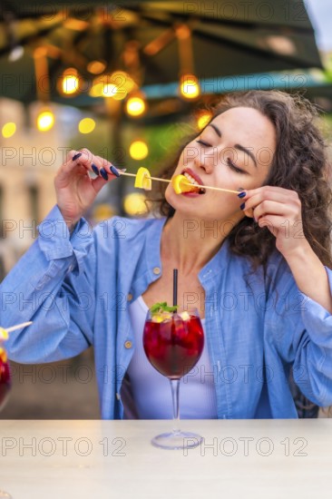 Vertical photo of a caucasian woman enjoying biting the citrus garnishing a cocktail in an outdoor terrace