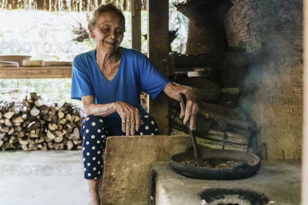 An elderly woman in a blue shirt sits by a rustic stove, stirring a frying pan with balinese coffee in a cozy kitchen. The natural light creates a warm, homely ambiance