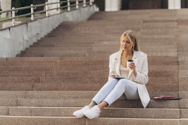 A businesswoman in casual business attire sits on outdoor steps, holding a coffee cup and using a tablet. A serene moment showcasing balance between work and relaxation