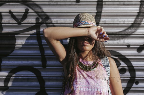 A stylish woman in a hat with chic accessories poses in front of a graffiti-covered metal door, exuding urban style and cool confidence, in bright natural light