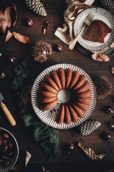 A delicious chestnut cake sits elegantly on a rustic table, surrounded by autumn leaves and pine cones. Its rich brown color reflects the cozy seasonal atmosphere perfectly