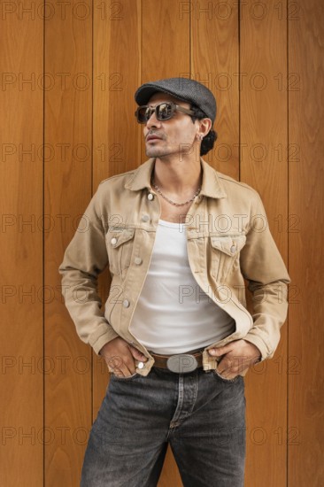 A fashionable man wearing sunglasses, a tan jacket, and a cap stands confidently against a wooden wall. His relaxed pose and stylish attire exude modern sophistication