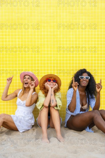 Three diverse women wearing summer clothes and hats are sitting on the sand, pointing upwards with their index fingers, in front of a yellow tiled wall
