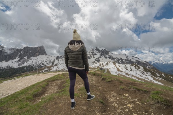 A woman stands at Passo Giau in the Dolomites, Italy, gazing at the snow-covered mountains under dramatic clouds. The rugged, scenic landscape showcases nature's beauty