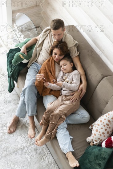 Top view image of a family of four celebrates Christmas in a warm, cozy setting. The mother, holding a child. The father, also seated, gently places his arm around both the mother and the child, in a serene, homely background with festive decorations