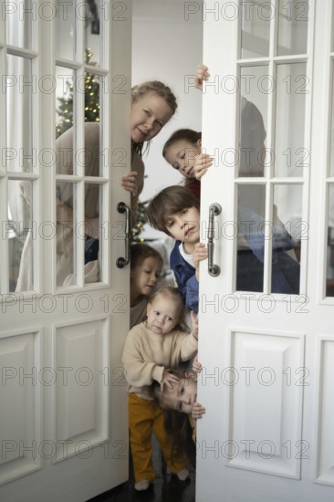 A cheerful family of five peeks around a white glass door, revealing bright smiles and curious faces In the background, a Christmas tree is subtly decorated with glowing lights
