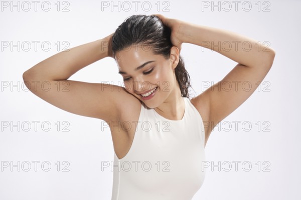 A female model in a white bodysuit enjoys a refreshing moment in a studio, her gaze looking down as she playfully puts her hair up, displaying a joyful demeanor against a plain background