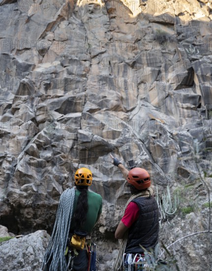 Two climbers equipped with helmets and ropes assess a towering rock face for a traditional climbing route in a natural setting, showcasing their adventurous spirit