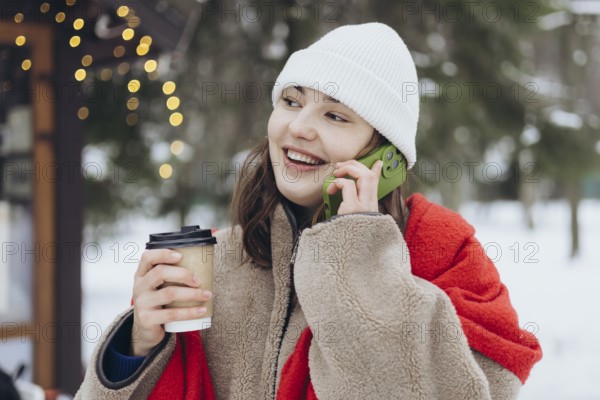 A young woman dressed in winter clothing holds a coffee cup and smiles while chatting on her phone outdoors on a snowy day, with soft-focus lights in the background