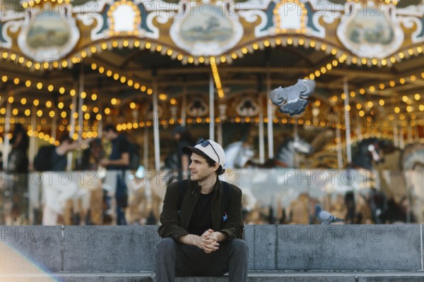 A young man sits casually in front of a brightly lit carousel in Brooklyn Bridge Park. Pigeons fly near him, adding dynamic motion to the scene. The festive atmosphere is captured beautifully