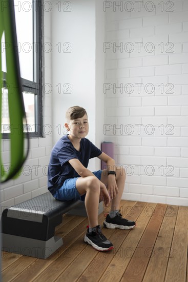 A young boy sits on a bench in a modern, brightly lit room with wooden floors and white brick walls, wearing casual athletic clothing and a smartwatch, looking thoughtful