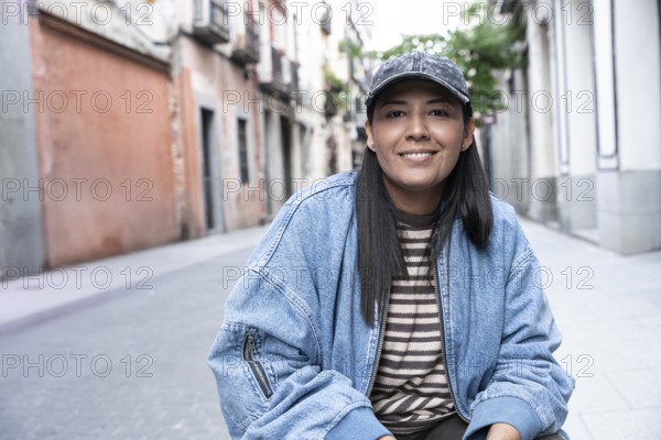 Latina woman wearing a striped shirt and denim jacket smiles confidently while sitting on a vibrant urban street lined with colorful buildings and lush greenery