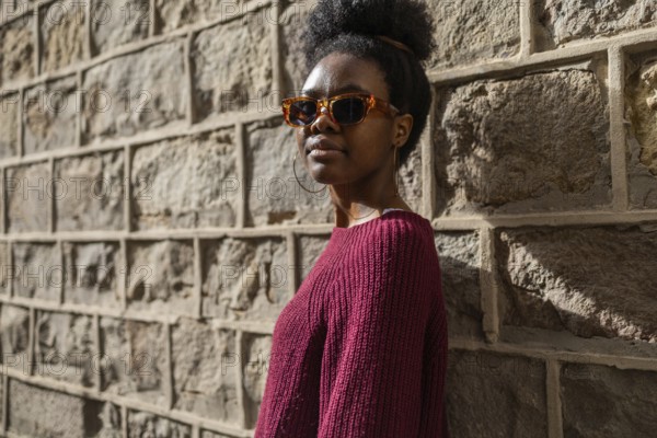 A stylish woman in a burgundy sweater wearing sunglasses stands confidently against a textured stone wall, casting a shadow in the warm sunlight of a clear day