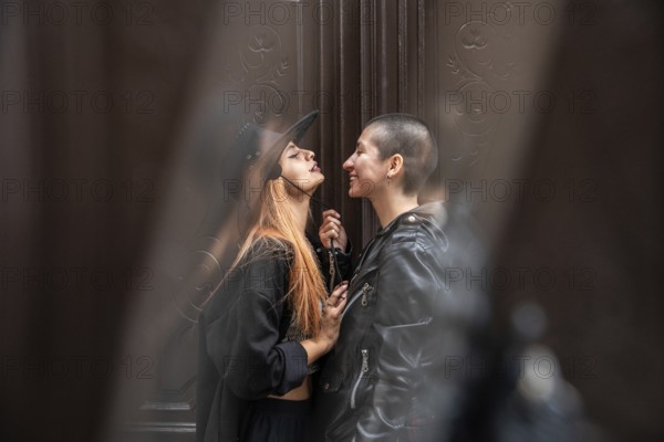 A lesbian couple sharing an intimate moment, surrounded by soft, muted surroundings. One partner in a hat leans forward, while the other smiles warmly, capturing their affection