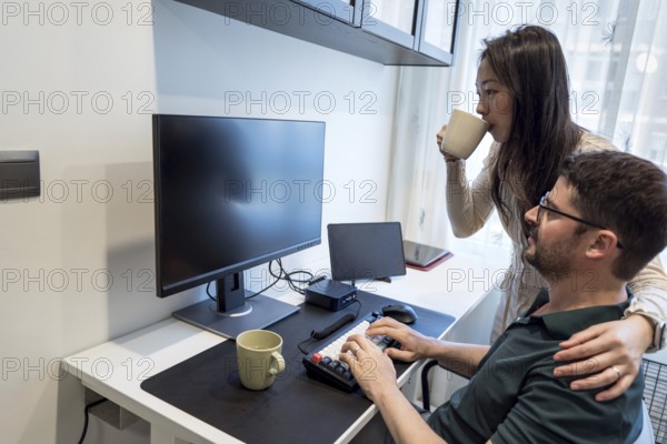 A diverse couple is sharing a cozy moment at home. One is working on a computer while the other enjoys a cup of coffee, embodying a relaxed and supportive atmosphere