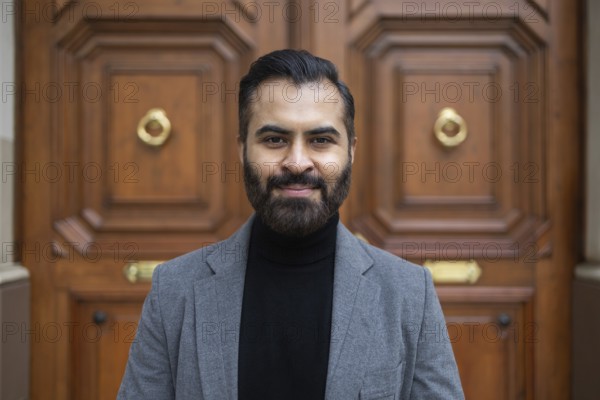 Portrait of a confident Indian businessman in a grey coat and black turtleneck, standing in front of a classic wooden door, looking directly at the camera