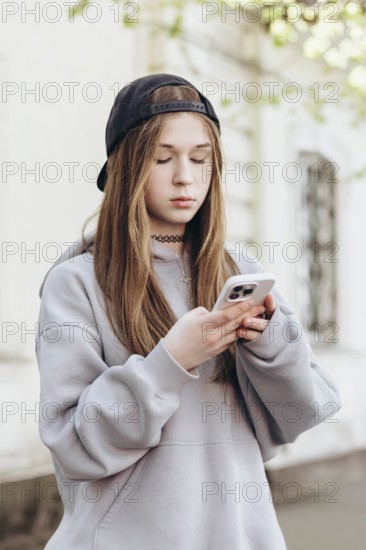 Teenage girl in casual clothing, engrossed in her smartphone while standing outdoors. She appears focused and engaged with the screen, possibly texting or browsing