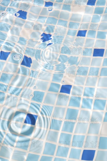 A close-up image of mosaic tiles under clear, rippling water, creating a serene and refreshing summer vibe. The blue and white tiles enhance the calming effect, with sunlight reflecting beautifully