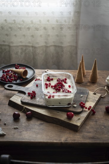 Rustic scene of homemade vanilla ice cream garnished with red currants and berries, served on a wooden board with scattered cherries