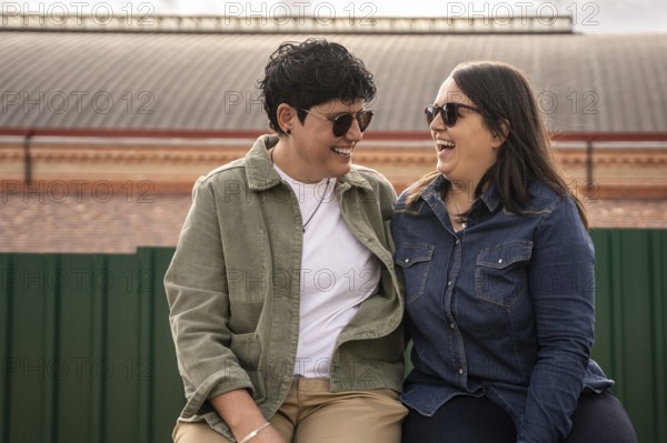A joyful curvy lesbian couple shares a light heart moment outdoors, sitting closely and wearing casual clothes. Their happiness and connection are beautifully captured