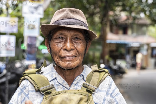 An elderly balinese man wearing a hat and a plaid shirt stands outdoors, looking into the camera with a serene expression. The background is a blurred street scene