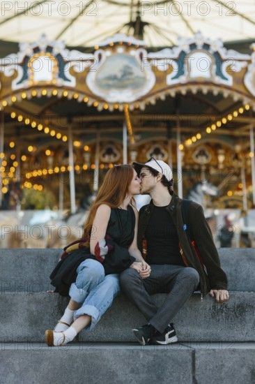 A couple shares a kiss on stone steps in front of a brightly lit carousel in Brooklyn Bridge Park. The atmosphere is romantic and vibrant, capturing the essence of a fun day at the fair