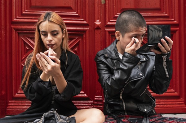 A lesbian couple sits together in front of a red door, each applying makeup with focus and care, highlighting a sense of individuality and shared moments