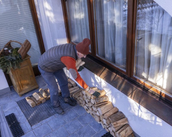 A senior man in casual winter wear stacks eco-friendly firewood next to his home on a clear day. The scene captures the essence of domestic life and sustainable living