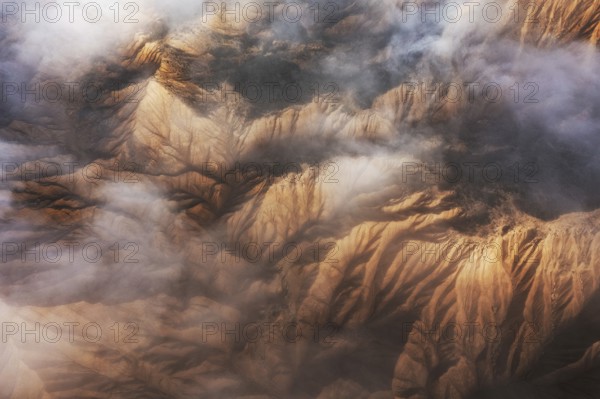 Captivating aerial view showcasing majestic mountain peaks partially enveloped in soft, swirling clouds The rugged terrain is highlighted by warm sunlight, creating a mesmerizing contrast