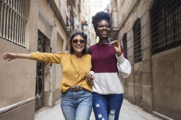 Two friends joyfully walk through a narrow street, one wearing sunglasses and the other holding them, capturing a moment of happiness and friendship on a sunny day