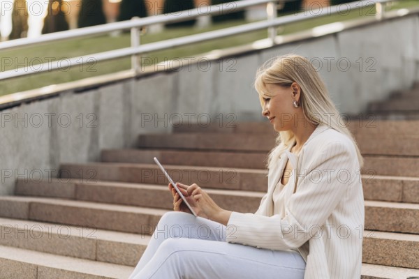 A businesswoman in casual business attire sits on outdoor steps while using a tablet. The setting has natural daylight, and the atmosphere is relaxed and focused