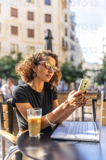 Vertical photo of a young Moroccan female freelancer working with mobile and laptop in a sidewalk cafeteria
