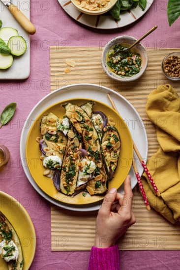 Top view of cropped unrecognizable hand serving a beautifully garnished roasted courgettes served on a vibrant yellow plate, alongside fresh ingredients and a savory dressing, showcasing a colorful and tempting presentation