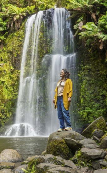 A woman stands calmly on a rock, surrounded by the lush greenery of a forest with a stunning waterfall cascading in the background, capturing a tranquil moment in the Great Ocean Road, Australia