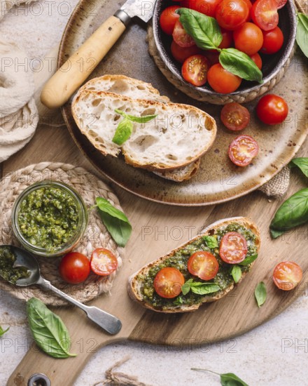 Rustic toasts topped with vibrant cherry tomatoes and fragrant pesto, presented on a wooden board. Fresh basil leaves and a knife add to the Mediterranean ambiance