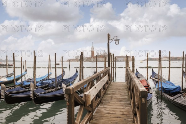Scenic view of a wooden dock leading to gondolas in Venice, Italy. The background showcases the iconic skyline and gentle clouds enhancing the serene atmosphere