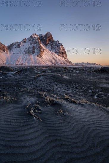 A stunning view of Stokksnes Mountain in Iceland, illuminated by the warm light of sunrise. The dark sand and icy terrain create a dramatic and serene winter scene