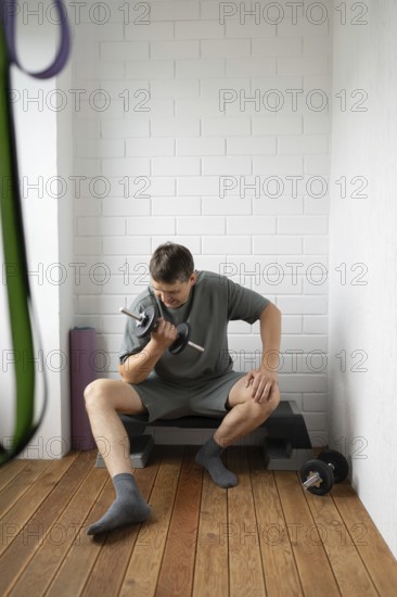 A man in comfortable clothing is seated on a step bench, focused on exercising with a dumbbell in a home gym setting, emphasizing strength training and fitness motivation