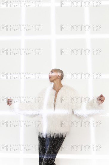 A middle-aged woman with a shaved head poses confidently in a chic white fur coat and stylish pants against a geometric background, embodying elegance and modern fashion