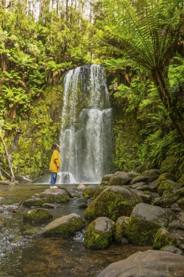 A woman stands by a waterfall in a lush forest, surrounded by moss covered rocks and vibrant ferns, offering a serene moment of connection with nature in the Great Ocean Road, Australia
