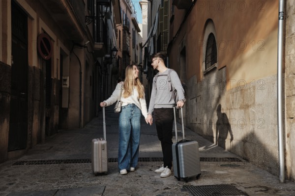 A young couple walks hand in hand down a narrow street, luggage in tow, as they search for their dream vacation home in the picturesque town of Calatrava, Majorca, Spain