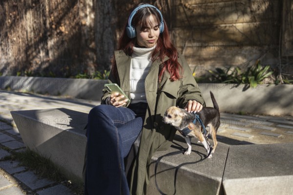 A young woman sits on a bench with headphones, holding a smartphone, while petting her small chihuahua outdoors on a sunny day. She appears relaxed and content