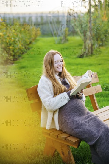 Beautiful pregnant woman with long red hair enjoys a serene moment on a park bench, reading a book. The lush green surroundings and her peaceful expression capture tranquility
