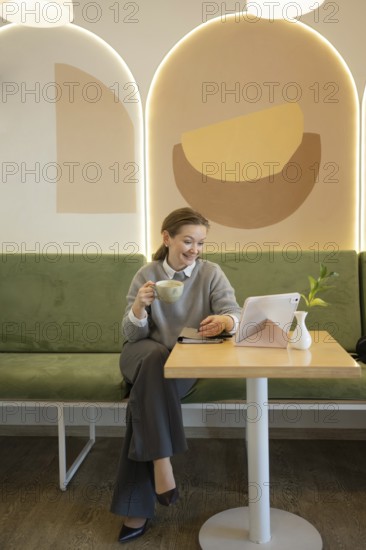 A woman relaxes in a stylish cafe, sipping coffee and using a tablet. The tranquil setting is enhanced by modern decor and soft lighting, creating a cozy ambiance
