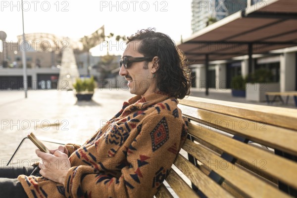 A man sits calmly on a bench outdoors with a smartphone Wearing sunglasses and a stylish patterned sweater, he enjoys the peaceful urban environment in the sunlight