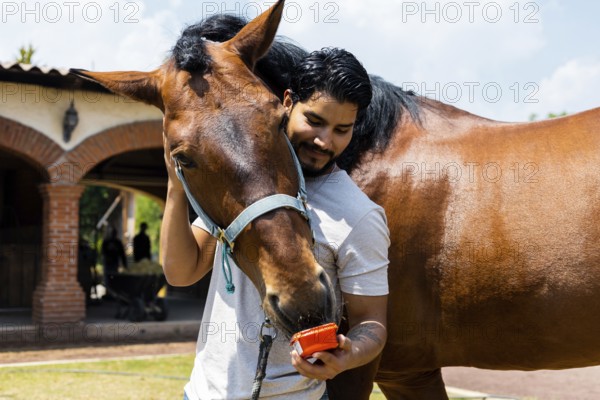 A man affectionately feeds his horse from a red container while embracing it on a sunny day at the ranch, showcasing a warm bond between humans and animals
