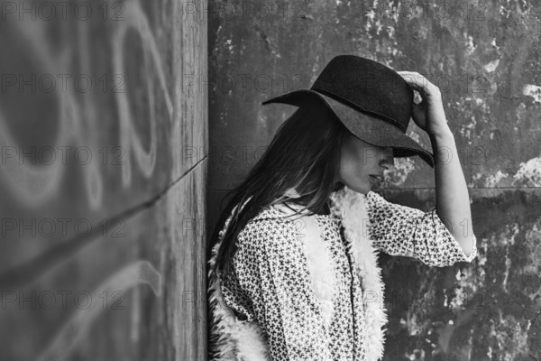 A black and white image of a woman in a hat resting against a textured wall. She wears a stylish outfit with a patterned top and fur vest, exuding a sense of calm and reflection