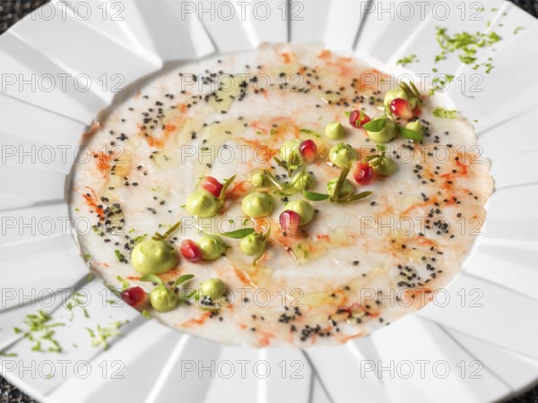 A beautifully arranged seafood carpaccio garnished with delicate green cream, red pomegranate seeds, and black sesame on a white plate. An artistic culinary presentation