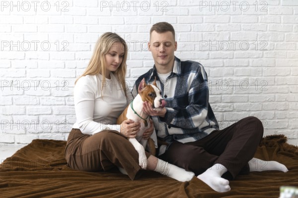 A young couple enjoys a peaceful moment with their Staffordshire Terrier puppy dog inside their cozy home, against a white brick wall background